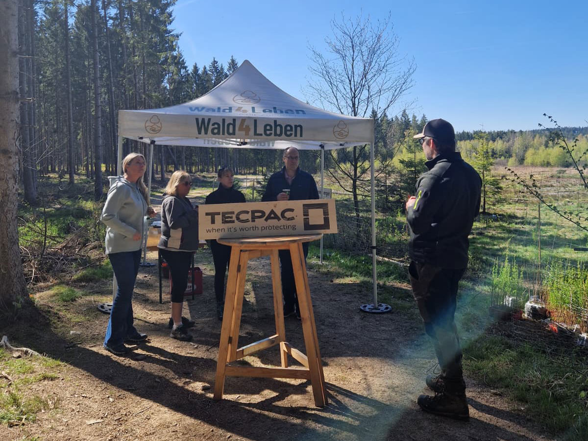 Several people under a "Wald 4 Leben" tent with a "TECPAC" sign in the forest.